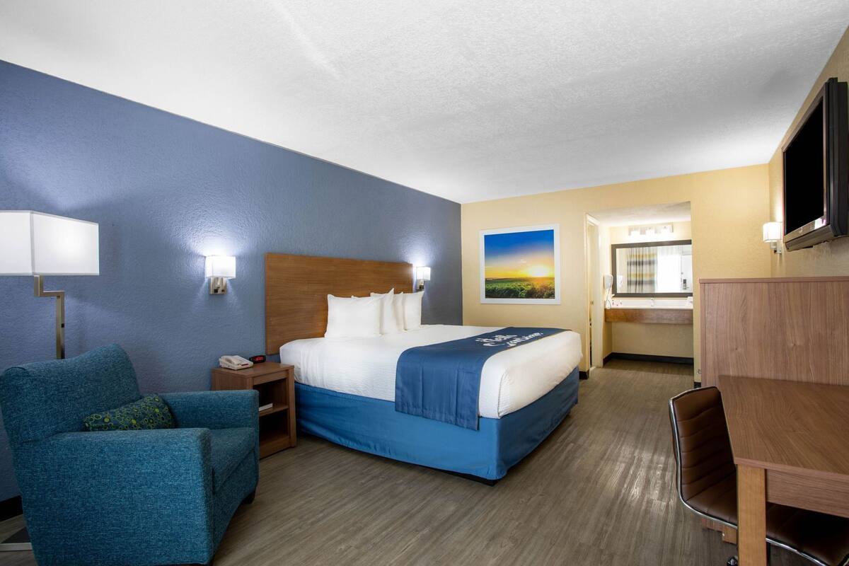 A hotel room with a bed, a desk, a chair, a nightstand, a lamp, a TV, and a painting on the wall.