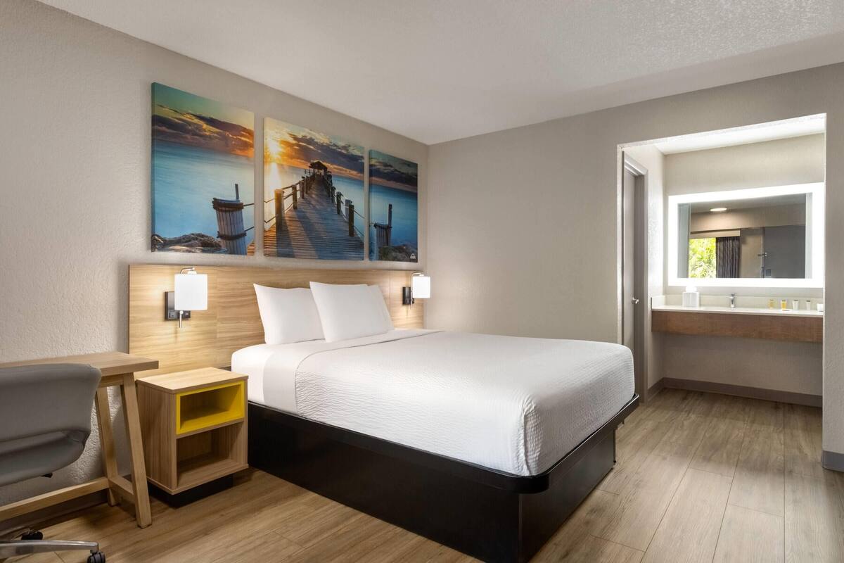 A hotel room with a bed, bedside tables, a desk, a chair, a bathroom with a sink, and wall art depicting a pier and sunset.