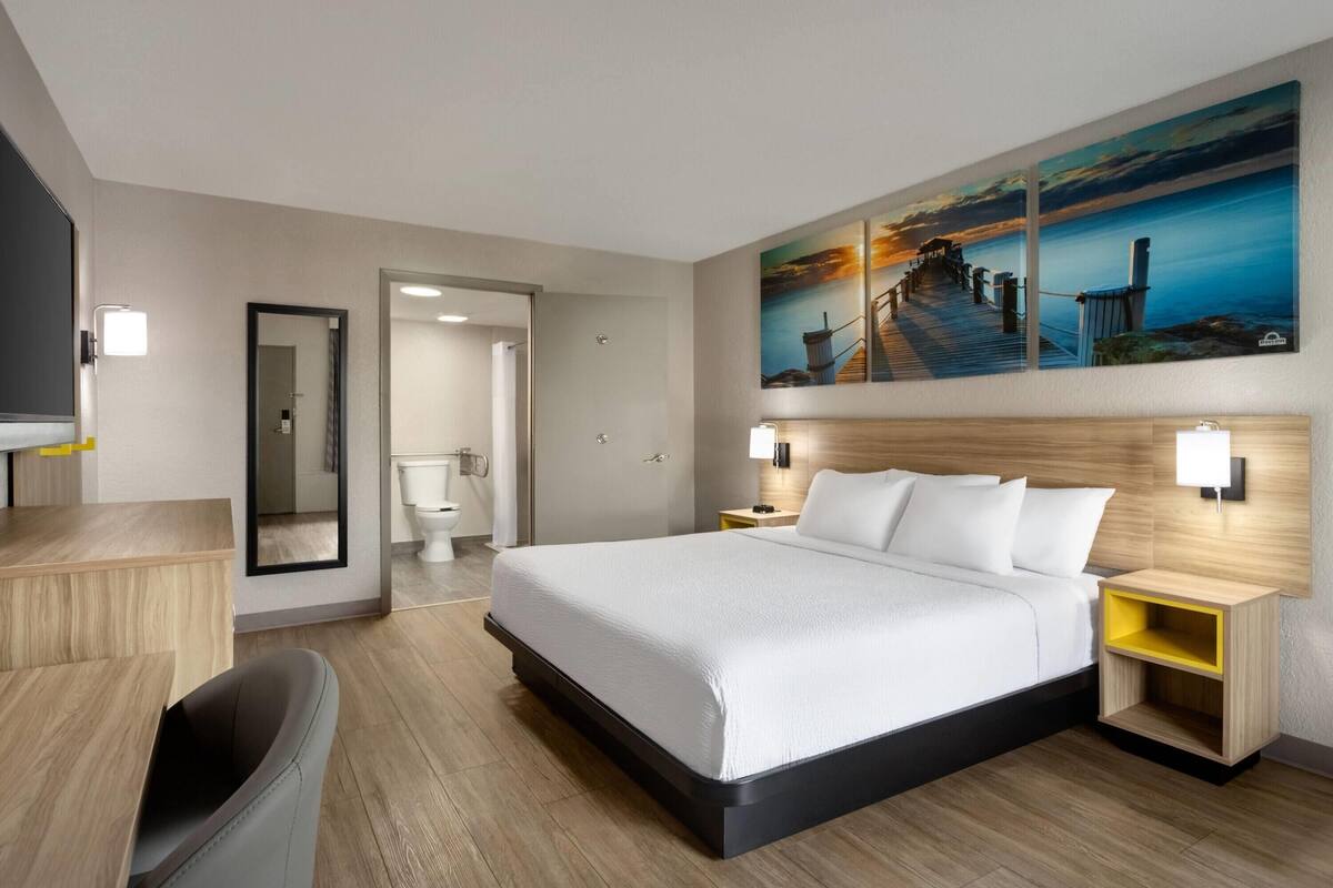 A modern hotel room with a large bed, bedside tables, a desk, and a chair. There is a bathroom visible through an open door.