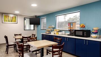 A kitchenette with a microwave, toaster, and various food items on the counter. There are wooden chairs and a table in the dining area.