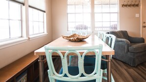 Un comedor con una mesa de madera, sillas azules y un cuenco tejido. Hay dos ventanas con persianas, un sofá gris y un perchero.
