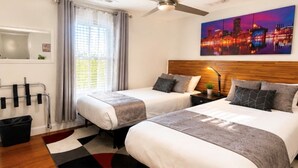 Habitació d'hotel amb dos llits, un quadre de paisatge urbà, un ventilador de sostre i una finestra amb cortines.