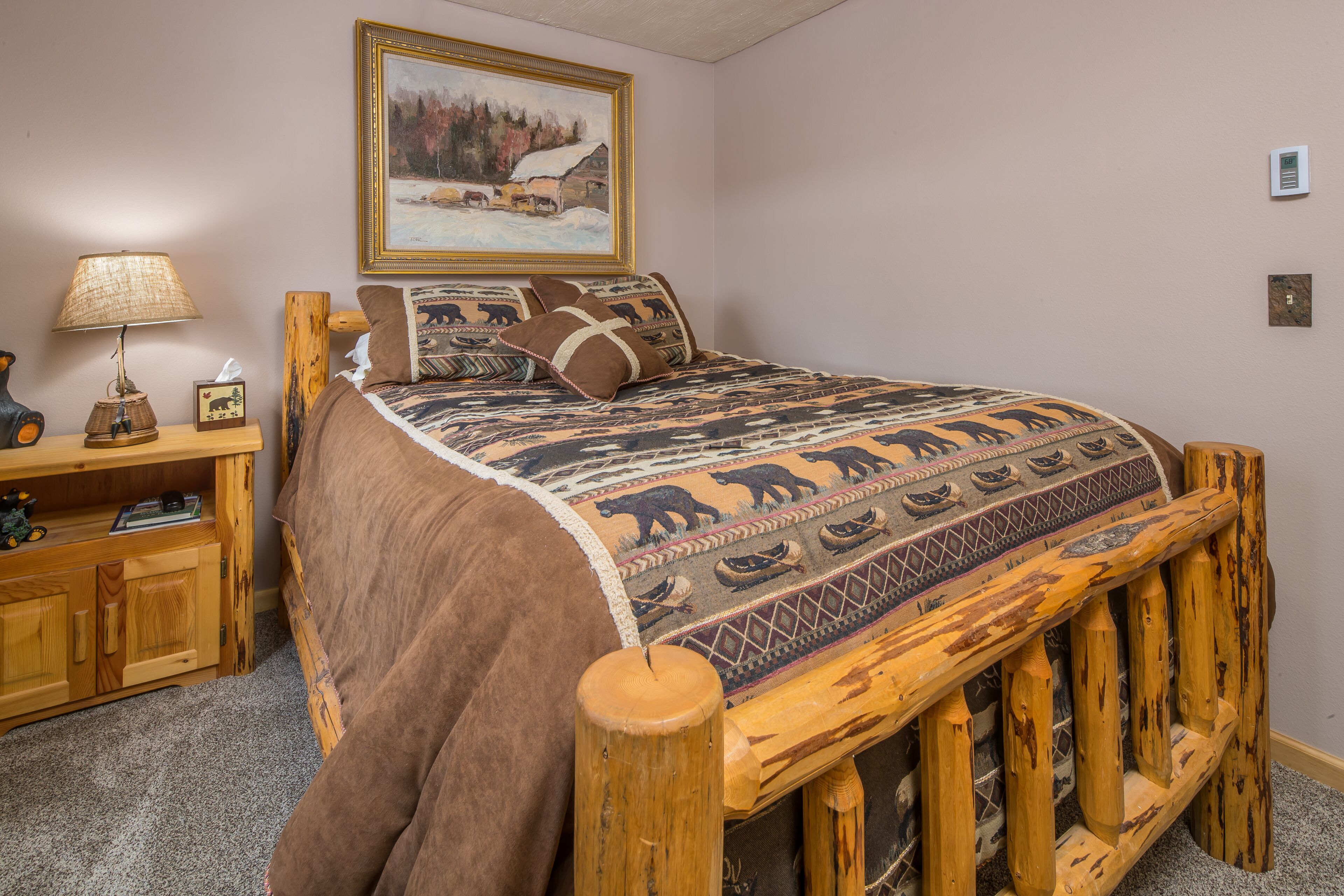 Ein hölzernes Bett mit einer Decke mit Bärenmotiv, ein Nachttisch mit Lampe und ein Gemälde einer verschneiten Landschaft an der Wand.