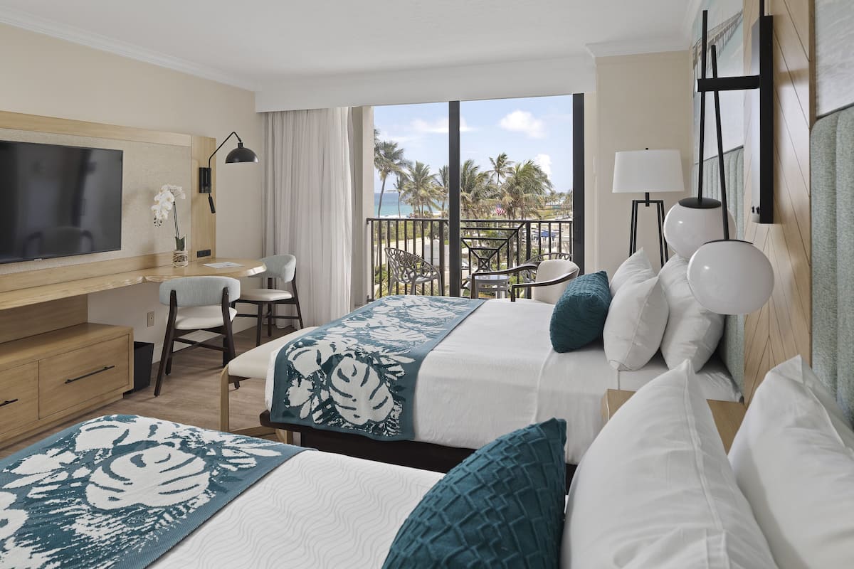 Habitación de hotel con dos camas, un escritorio, un televisor y un balcón con vista a la playa y palmeras.