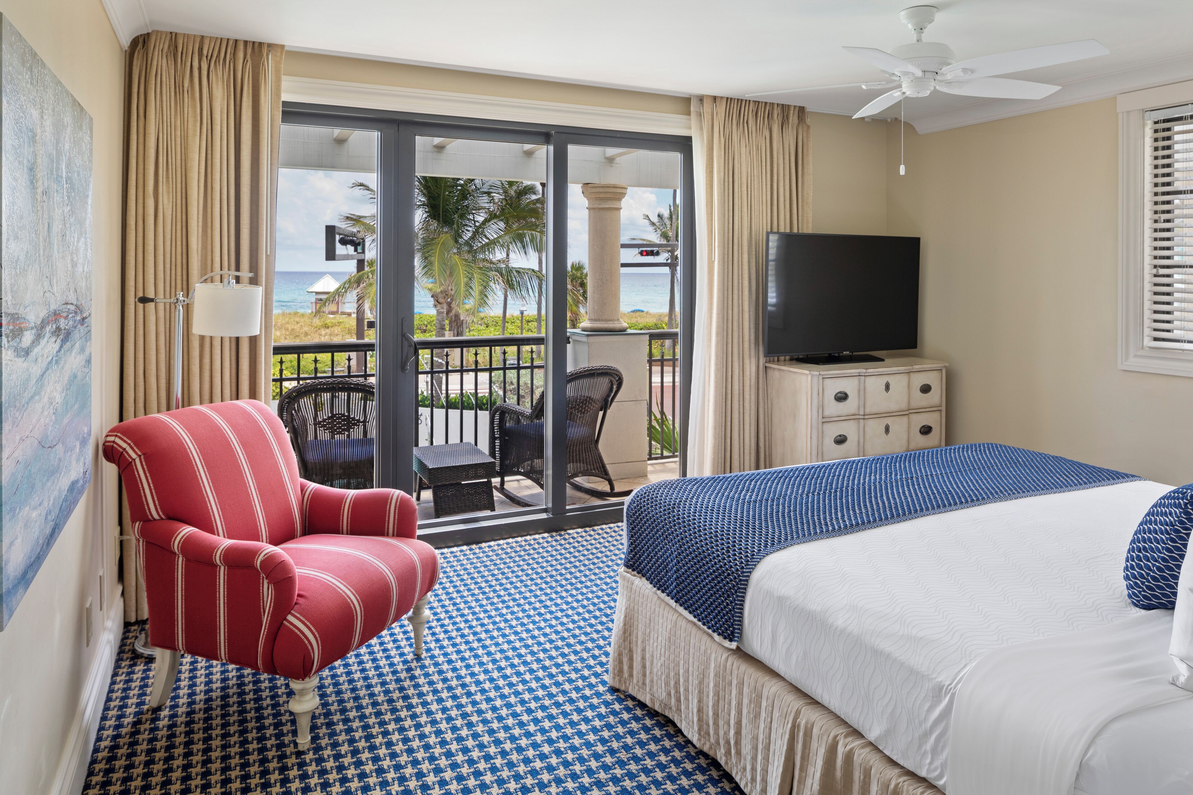 A hotel room with a bed, a TV, a red armchair, and a balcony with a view of the ocean.