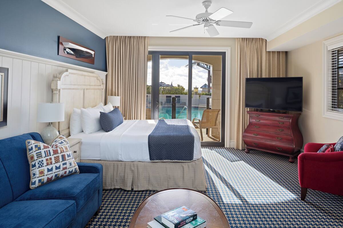 Habitación de hotel con cama, sofá azul, sillón rojo, televisión y balcón con piscina.