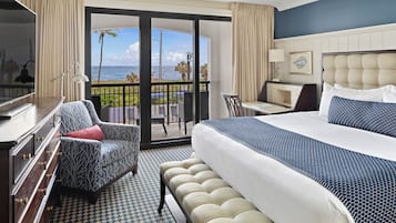 A hotel room with a large bed, a desk, a chair, and a TV. The room has a view of the ocean and palm trees.
