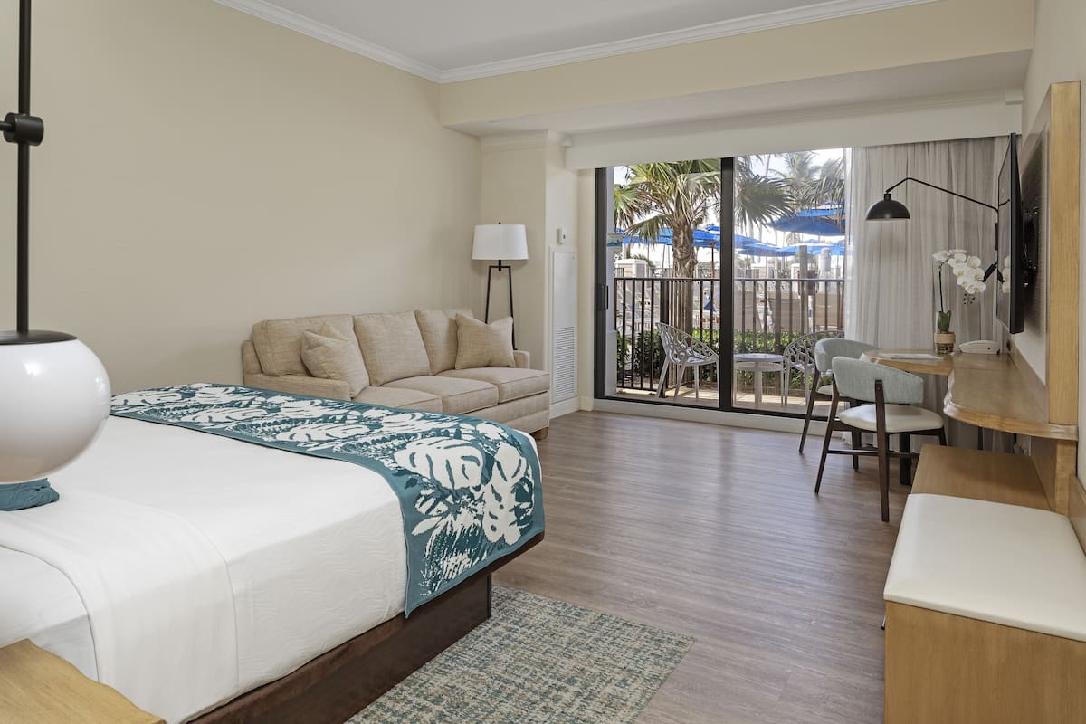 Habitación de hotel con cama, sofá, escritorio y silla, televisor y balcón con mobiliario exterior.