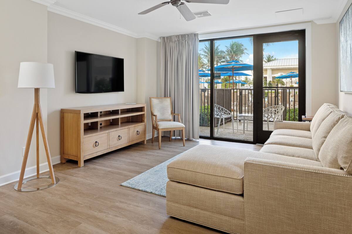 Una sala de estar moderna con televisor de pantalla plana, mueble de entretenimiento de madera, sofá color beige y vista al área exterior con sombrillas y palmeras.