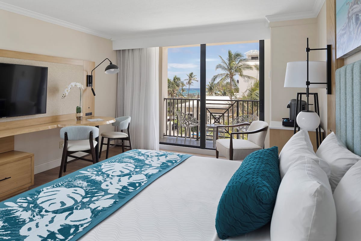 Habitación de hotel con una cama grande, un escritorio con televisor, un balcón con sillas y vistas a la playa.