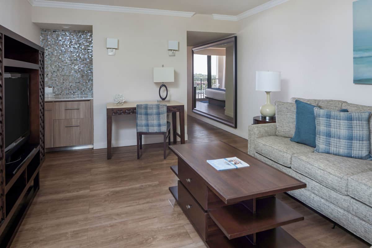 Una sala de estar con una mesa de centro de madera, un sofá con cojines azules, un televisor, un escritorio con una silla y una zona de cocina con salpicadero de azulejos.