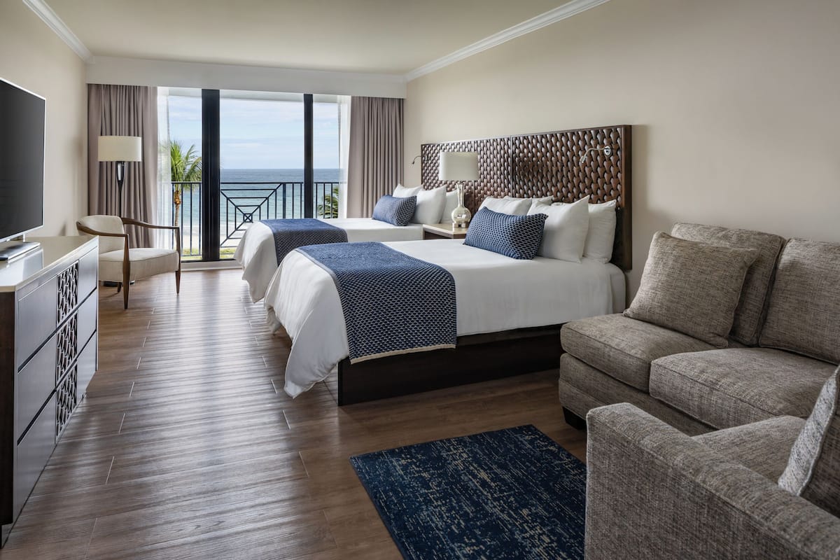 Habitación de hotel con dos camas, un sofá, televisión y balcón con vista al océano.
