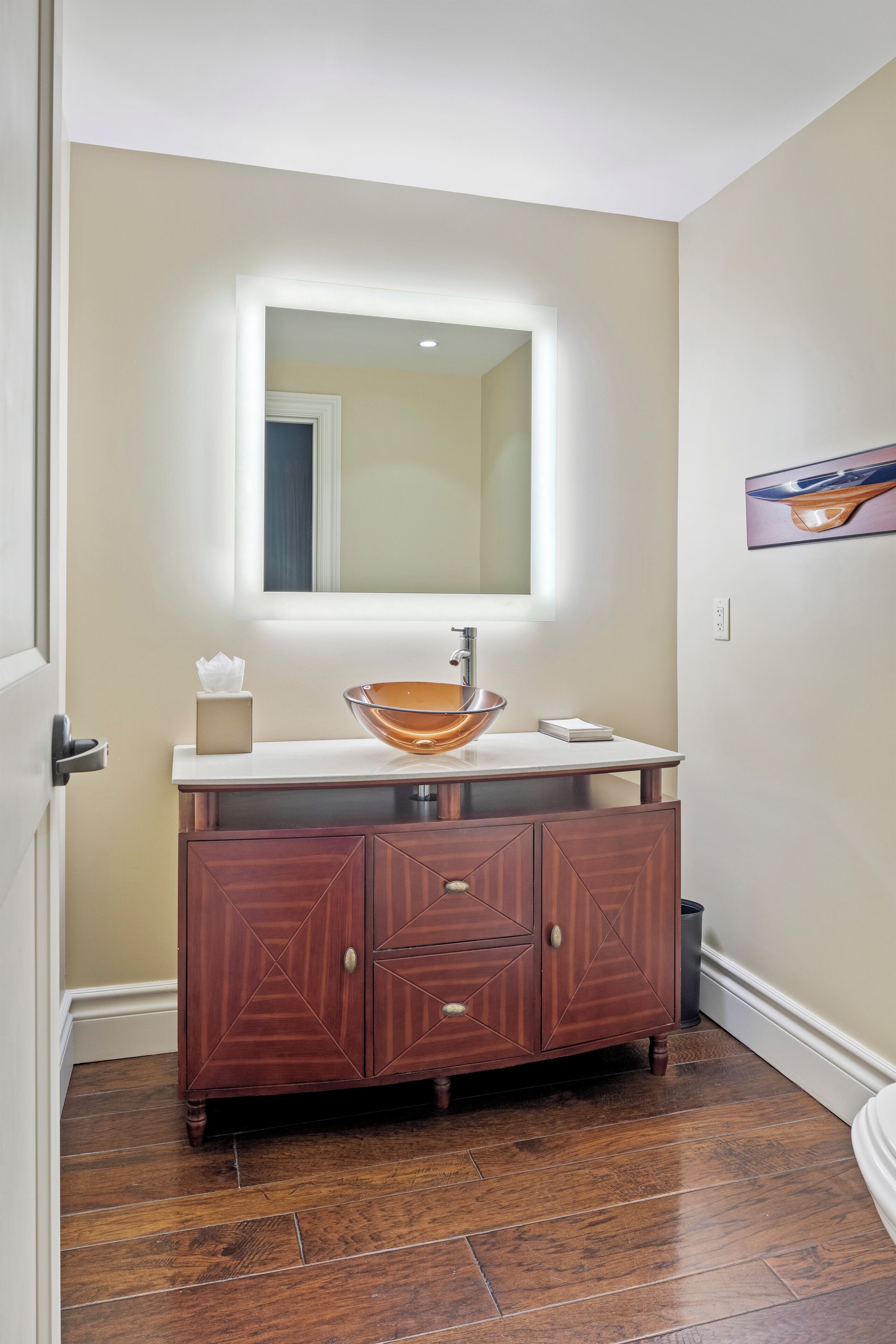 A bathroom with a wooden vanity, a glass bowl sink, and a mirror with a light above it.
