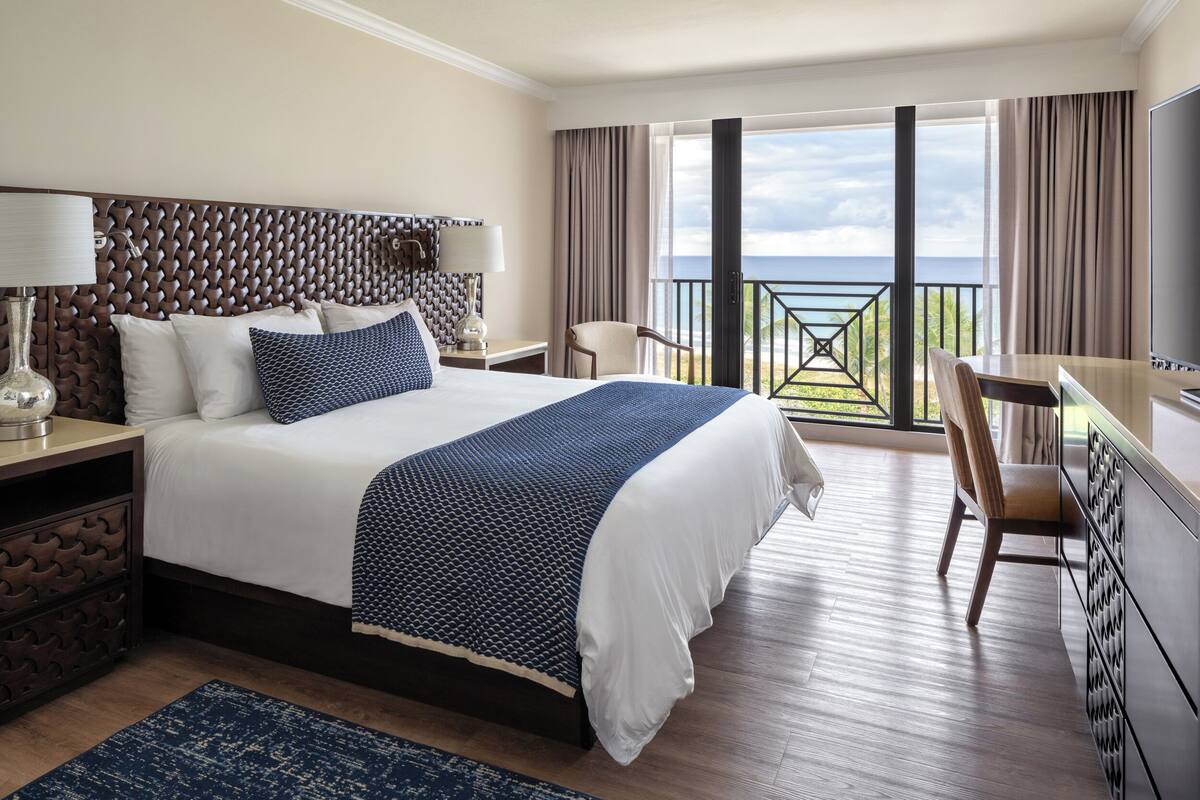Habitación de hotel con una cama grande, un escritorio, una silla y un balcón con vistas al mar.