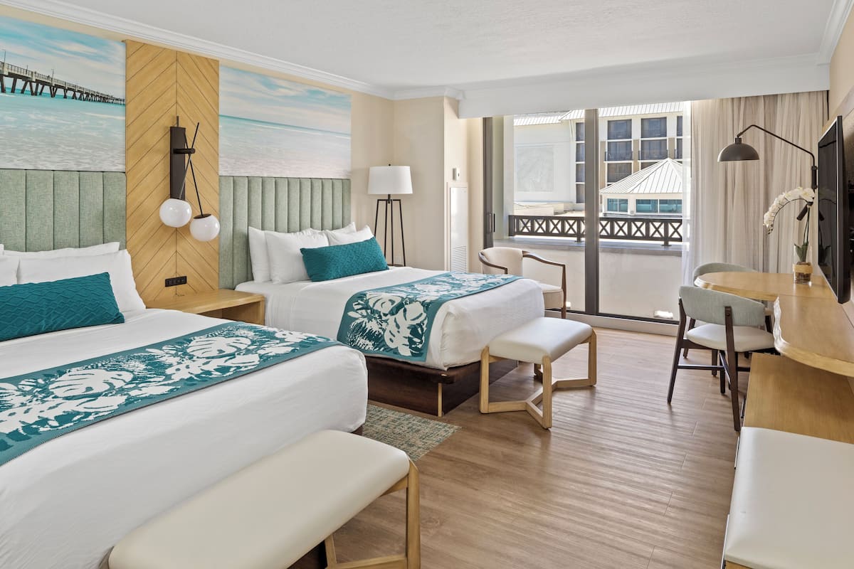 Habitación de hotel con dos camas, un escritorio, una silla y vistas a un edificio a través de amplios ventanales.