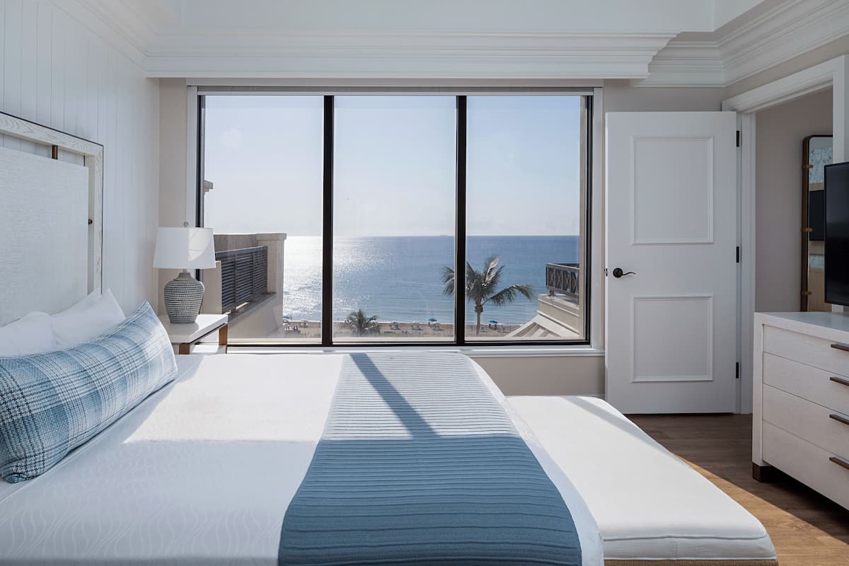 Un dormitorio con un amplio ventanal con vistas al mar, una cama con colcha azul a rayas, una mesita de noche con una lámpara y un televisor sobre un aparador.