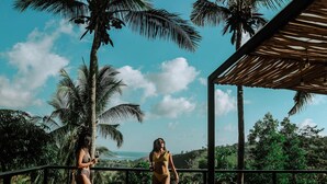 A balcony with two women in bikinis, palm trees, and a view of the ocean.