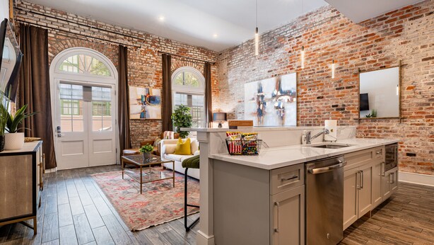 A modern kitchen with a brick wall, a large island with a sink, and a dining area with a sofa and table. - Luxury Condo Walk Anywhere & Everywhere! The Bordeaux (New Orleans)
