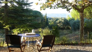 A table with a patterned tablecloth and two chairs, set on a gravel surface with a view of a forested landscape.