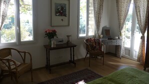A room with a bed, a desk, a chair, a table with flowers, and a painting on the wall.