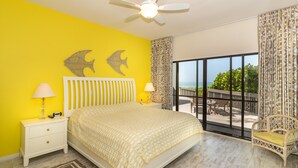 Une chambre à coucher aux murs jaune vif, avec un lit, deux décorations murales représentant des poissons, un ventilateur de plafond et une vue sur l’extérieur grâce à de grandes fenêtres.