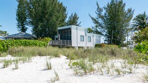 Une maison de plage dotée d’une terrasse, entourée de sable et de végétation.