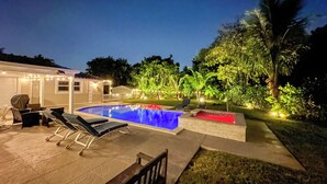 Una piscina con sillas de descanso, un porche cubierto y un jardín bien iluminado con árboles y plantas.