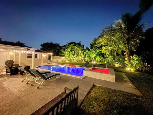 Une piscine avec des chaises longues, un patio couvert et un jardin bien éclairé, agrémenté d’arbres et de plantes.