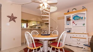 A dining area with a white table and chairs, a ceiling fan, and a kitchen in the background. - Ocean Park Terrace 202 · Ocean Park 202 · Rooftop Deck · 1-Month Min (Bradenton Beach)