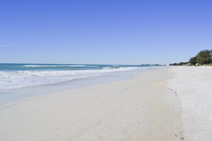 A wide sandy beach with gentle waves, a clear blue sky, and a line of trees in the distance. - Ocean Park Terrace 202 · Ocean Park 202 · Rooftop Deck · 1-Month Min (Bradenton Beach)