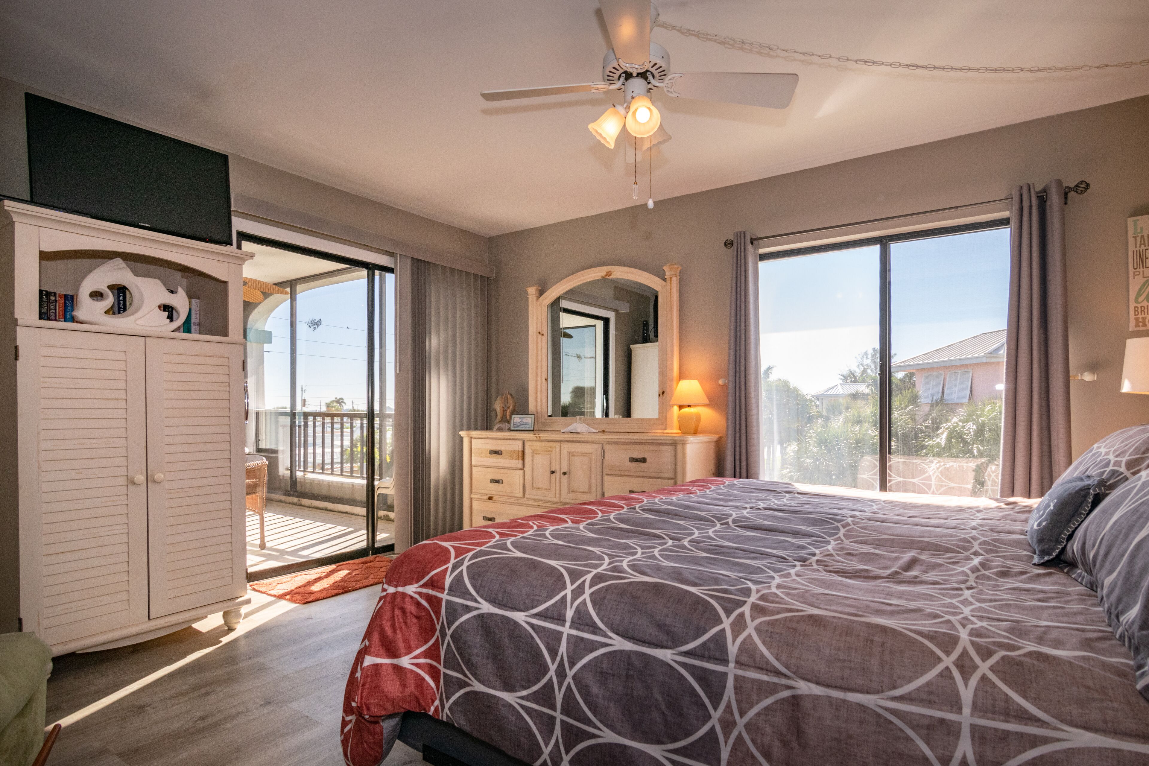 A bedroom with a large bed, a ceiling fan, a dresser, a mirror, and a balcony with a view.