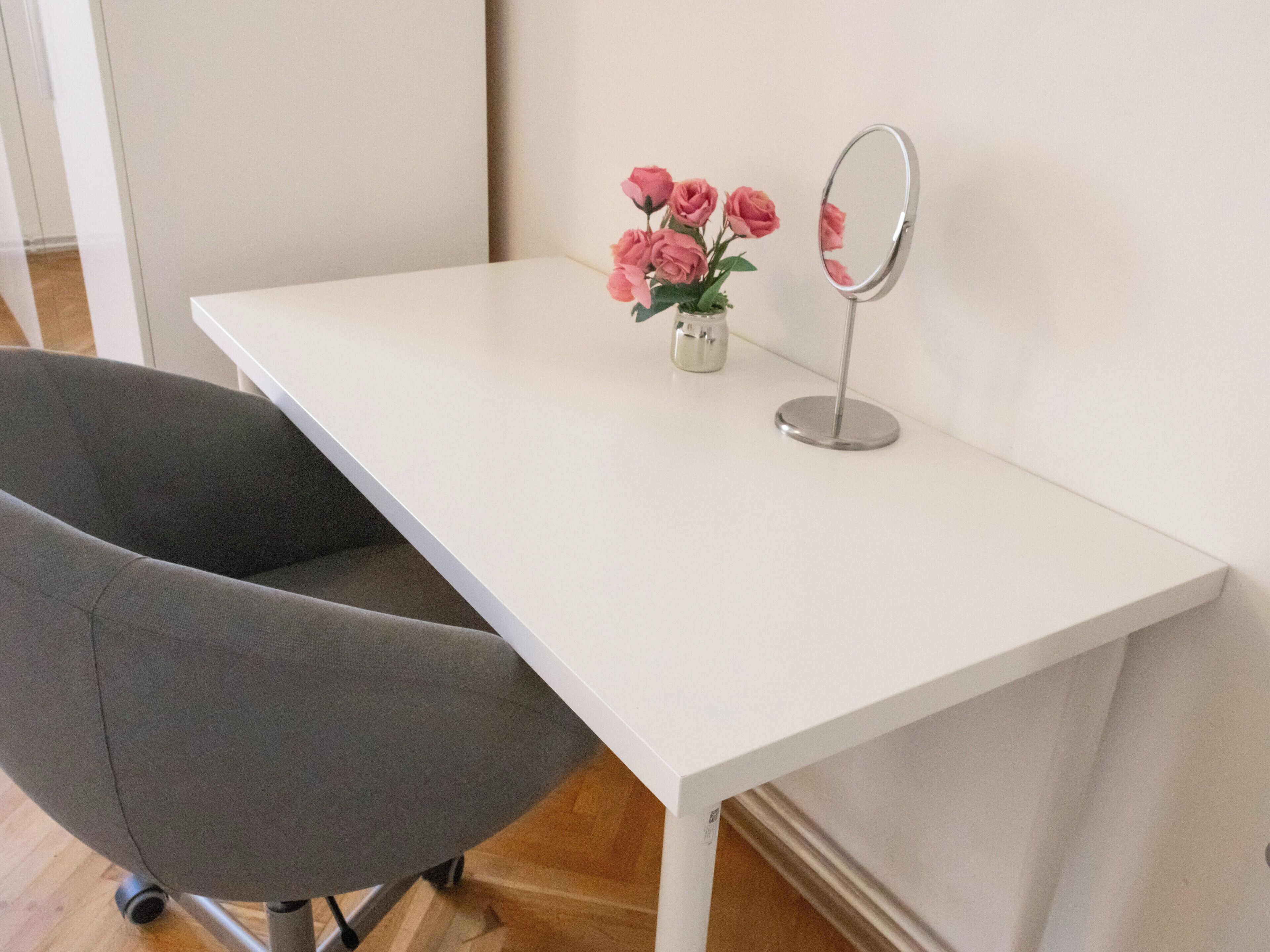 A white desk with a grey chair, a small vase with pink flowers, and a mirror.