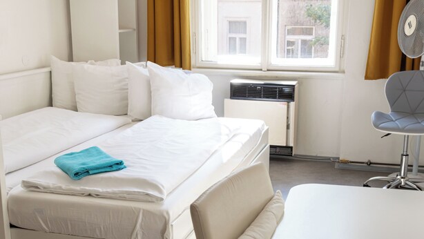 Cama bem arrumada com lençóis brancos e toalha azul, uma mesa pequena, uma cadeira e uma janela com cortinas.