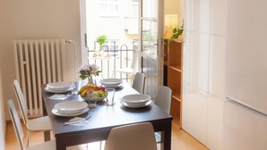 Uma área de refeições com uma mesa posta para quatro pessoas, cadeiras, uma tigela de frutas e uma janela com vista para a rua.