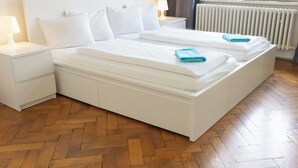 Quarto de hotel com duas camas, mesa de cabeceira branca com luminária, radiador e janela com cortinas.