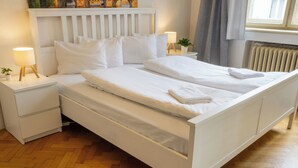 Een netjes opgemaakt bed met witte lakens en kussens, een nachtkastje met een lamp en een potplant op het nachtkastje.