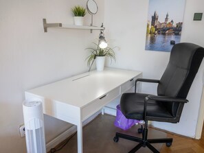 A modern office space with a white desk, a black office chair, a small potted plant, a wall-mounted mirror, and a framed cityscape picture.