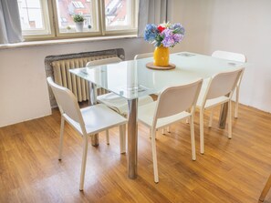 A modern dining area with a glass table and white chairs, a yellow vase with flowers, and a view of buildings through a window.