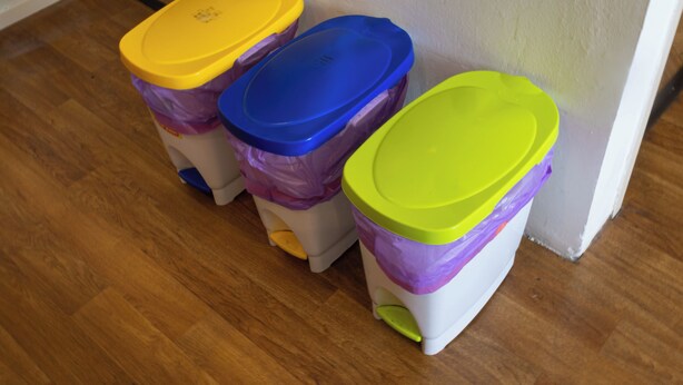 Drie afvalbakken met deksels in verschillende kleuren staan tegen een muur.