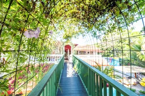A covered walkway with a green railing, surrounded by lush greenery and a red building in the distance. - 🌴16 Bed Compound Oasis 🌴Downtown Jaco with Private Pool & Hot Tub (Jaco Beach)