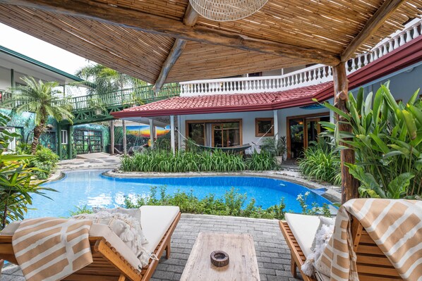 A poolside area with lounge chairs, a wooden table, and a red-roofed building.