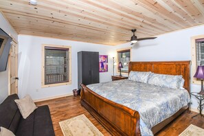 A bedroom with a wooden ceiling, a large bed with a patterned bedspread, a dark sofa, and a small table with a lamp. - Woods-stock (McHenry)