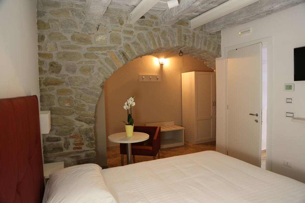 Une chambre avec une arche en pierre, un lit, une petite table sur laquelle est posé un vase, une chaise et une armoire.