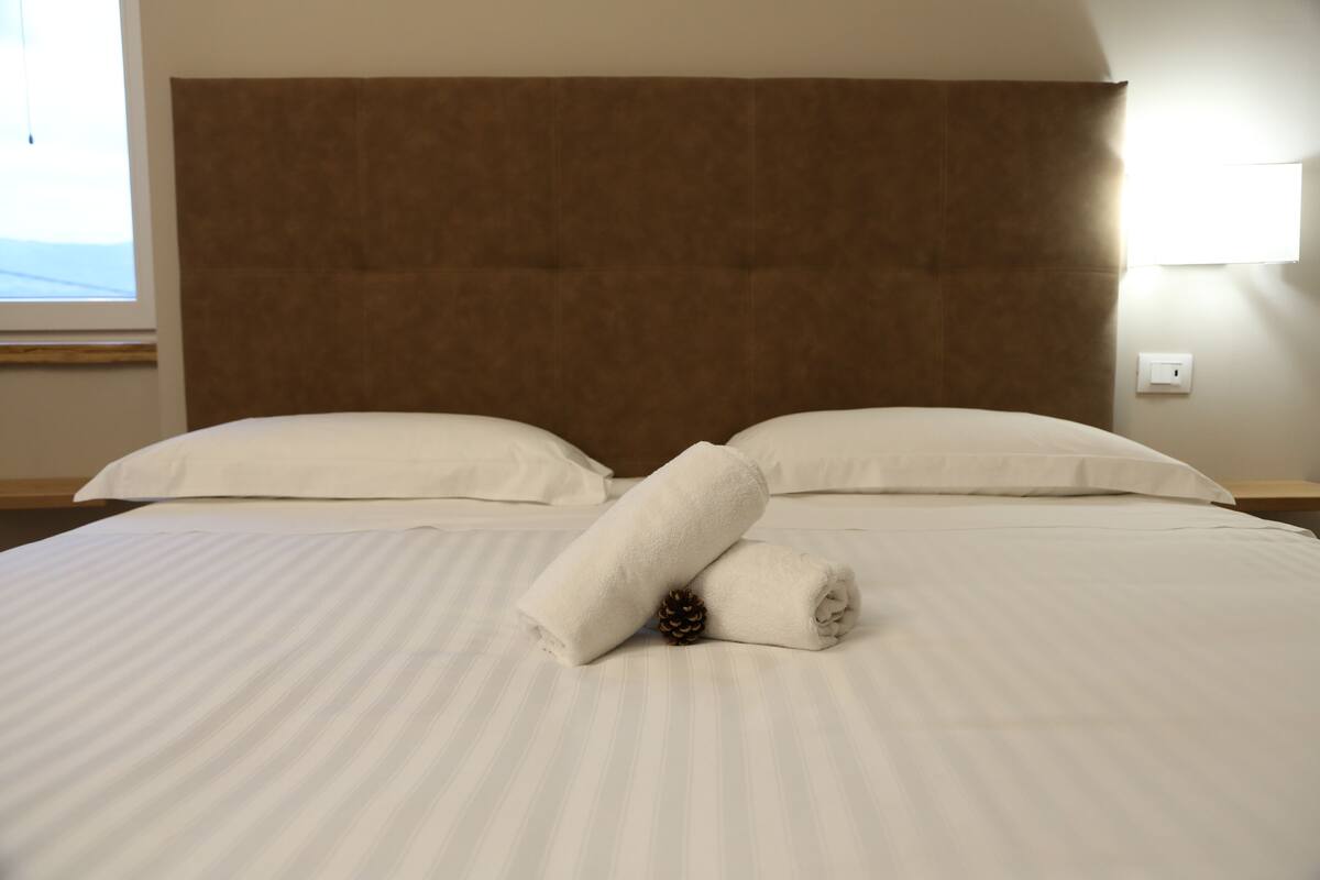 Un lit bien fait, avec du linge de lit blanc, une tête de lit marron et un centre de table décoré d’un pommeau de pin.