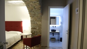 A bedroom with a red headboard, a small round table, and a red chair. There is a doorway leading to a bathroom with a sink and a mirror. - Montis (San Massimo)