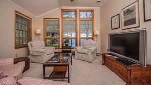 A living room with two beige armchairs, a glass coffee table, a flat-screen TV, and large windows with wooden frames. - Paradise Found (Oakland)