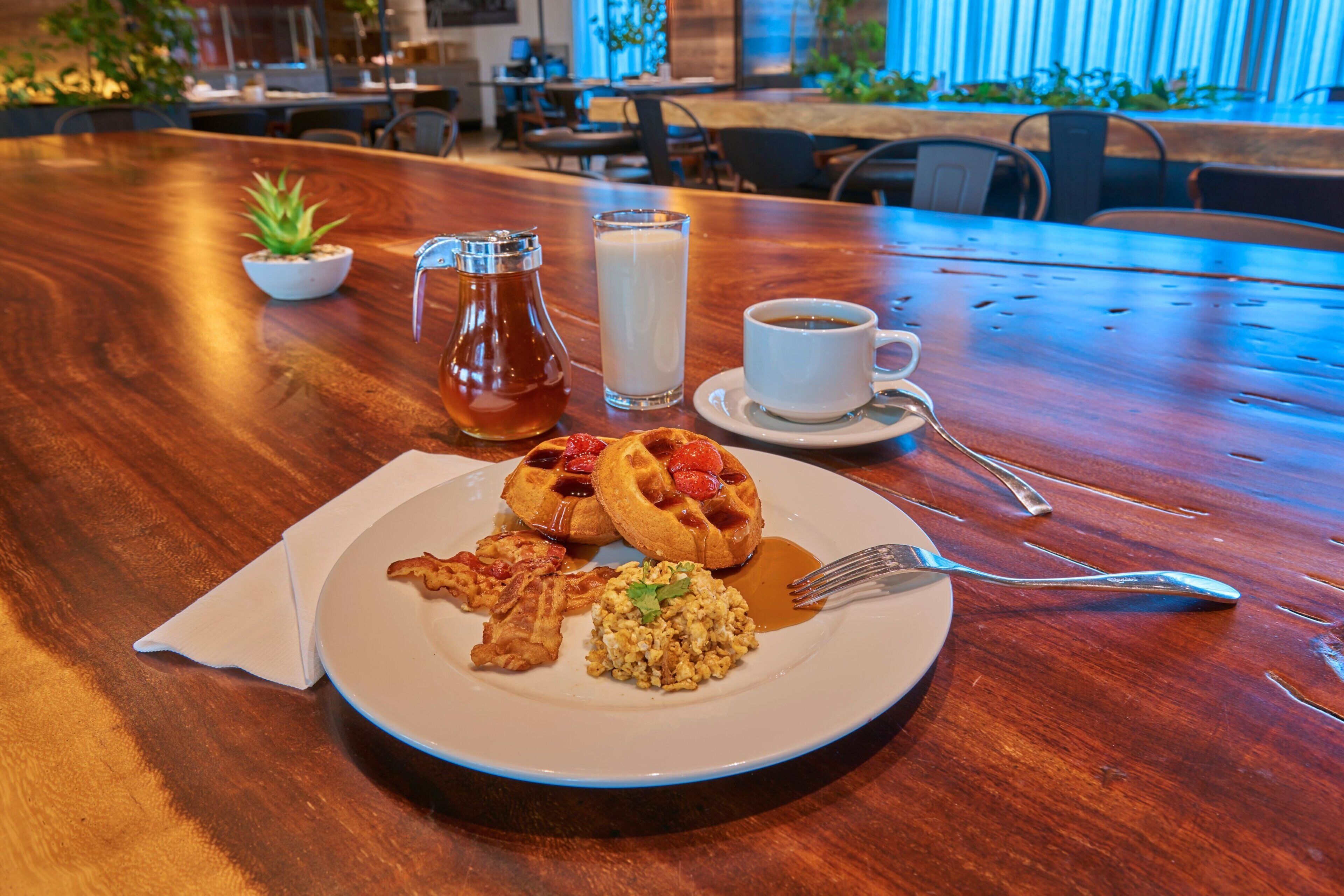 A breakfast setting with waffles, scrambled eggs, and syrup on a plate, a glass of milk, and a cup of coffee on a wooden table.