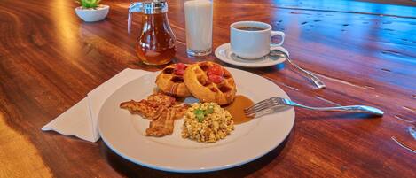 A breakfast setting with waffles, scrambled eggs, and syrup on a plate, a glass of milk, and a cup of coffee on a wooden table.