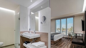 Habitación de hotel moderna con una cama grande, un escritorio, una silla, un baño con lavamanos y espejo, y vistas de la ciudad a través de amplias ventanas.