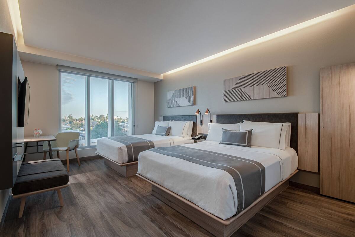 Ein modernes Hotelzimmer mit zwei Betten, einem Schreibtisch und einem großen Fenster mit Blick auf die Stadt.